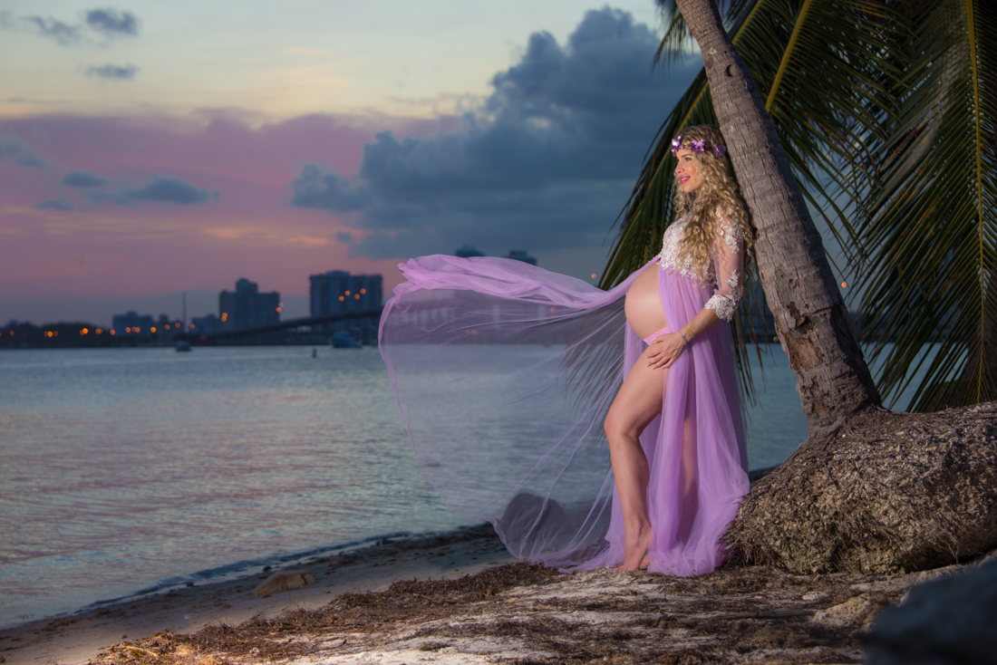 mom to be leaning on palm tree with floating dress at the beach miami skyline sunset