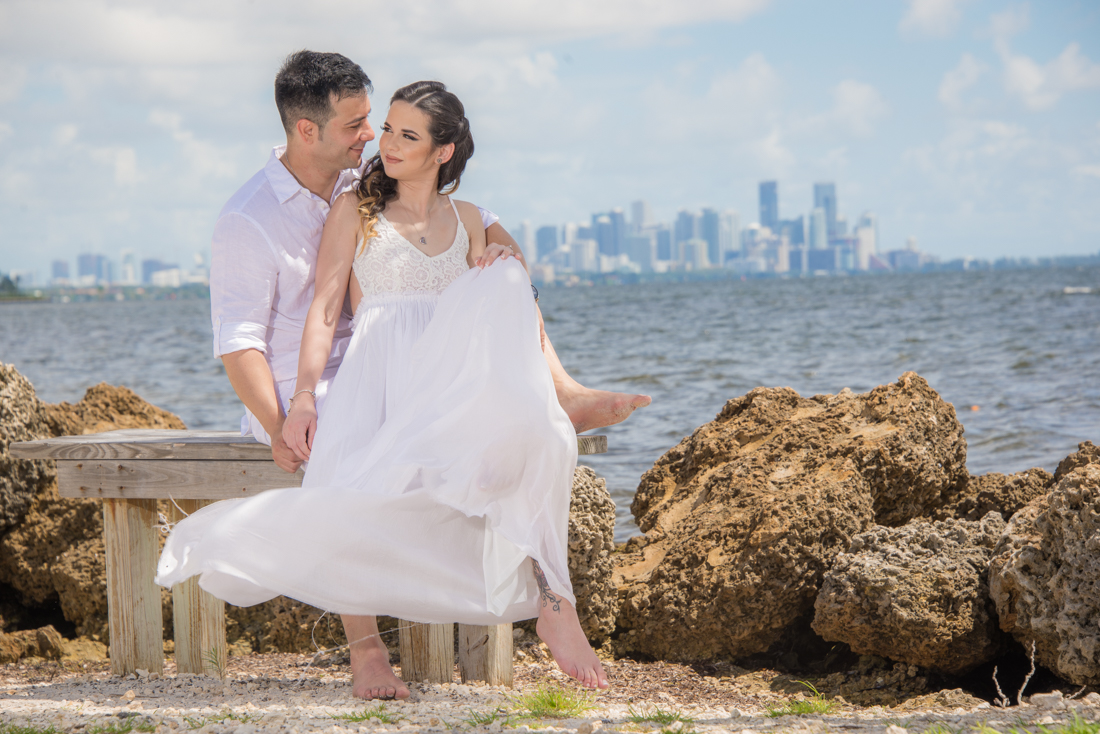 engagement couple picture starring at each other sitting on bench by the beach